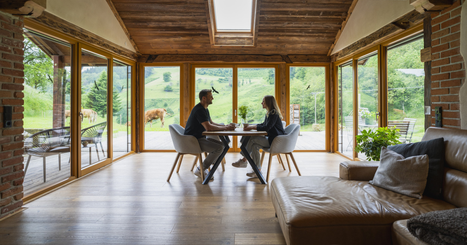 Im Wohnzimmer der Alten Backstube sitzt ein junges Paar am Esstisch. Im Hintergrund ist das satte grün der umliegenden Wiesen mit Rindern zu sehen.