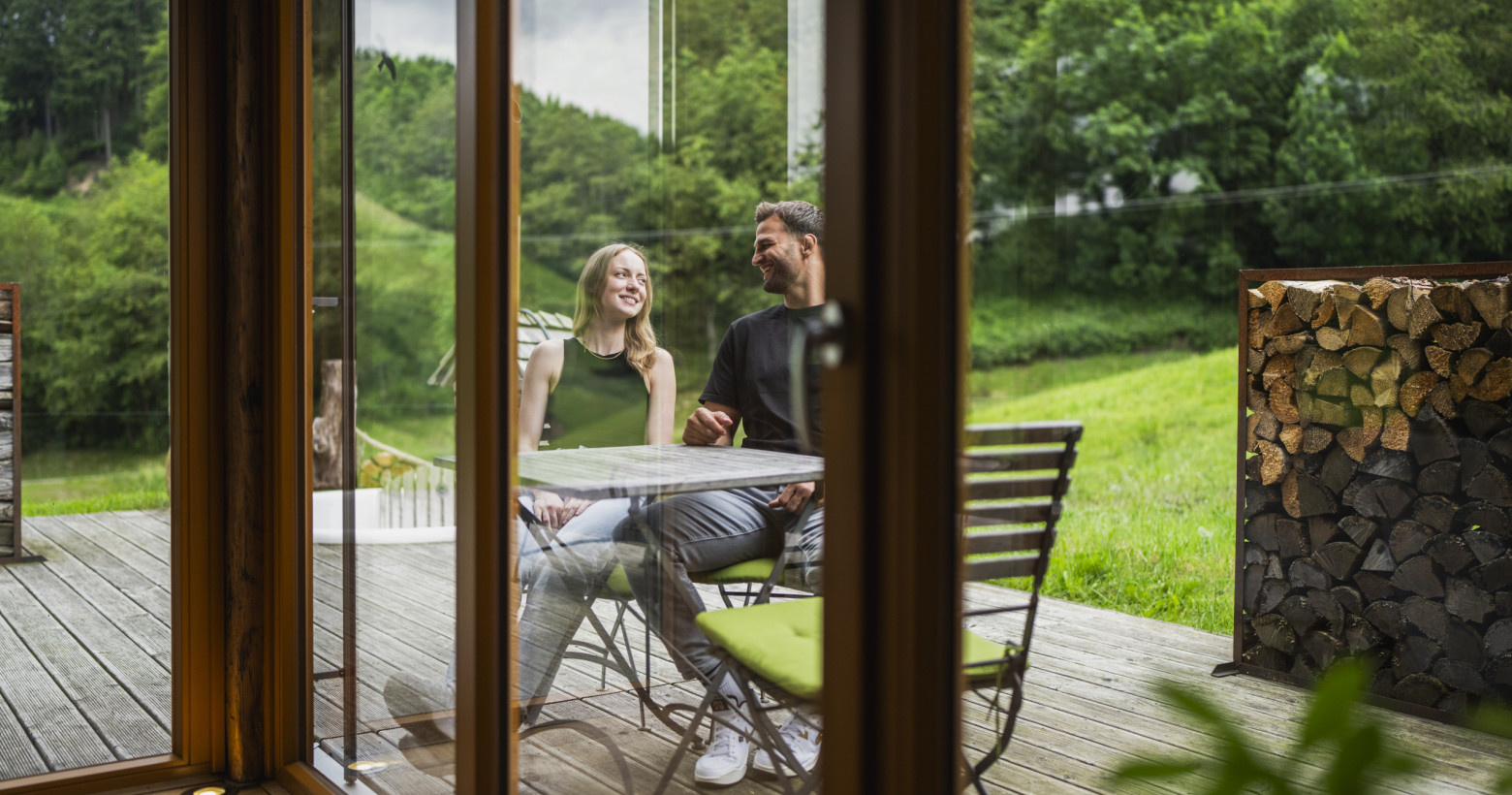 Durch die Scheibe ist ein junges Paar auf der Terrasse zu sehen. Sie sitzen am Tisch und unterhalten sich.