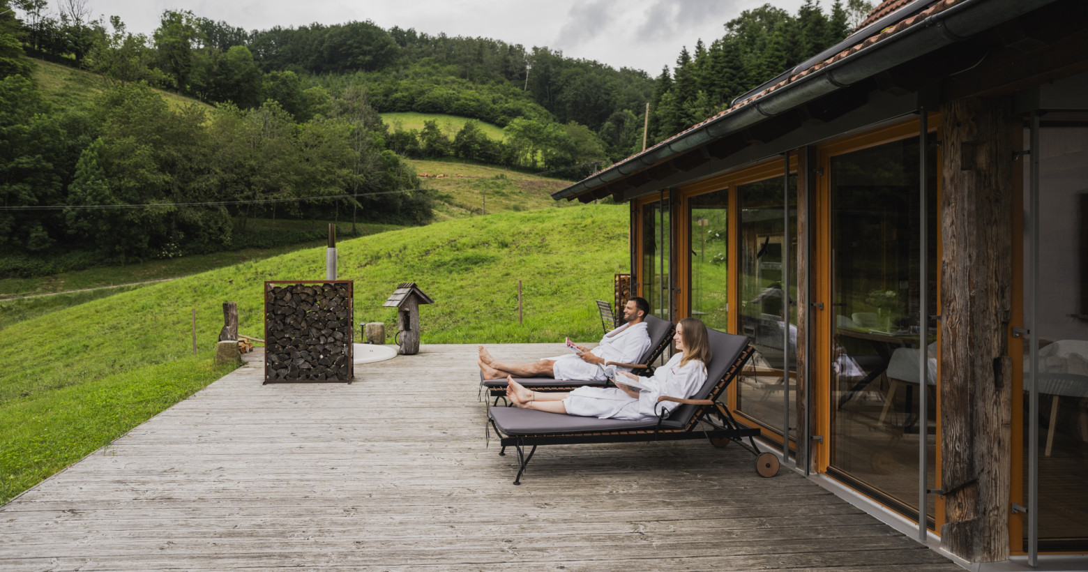 Auf der Terrasse liegt ein junges Paar in den Bademänteln auf den Liegestühlen und liest. Im Hintergrund sind die saftigen Wiesen und ein Stück Schwarzwald zu sehen.